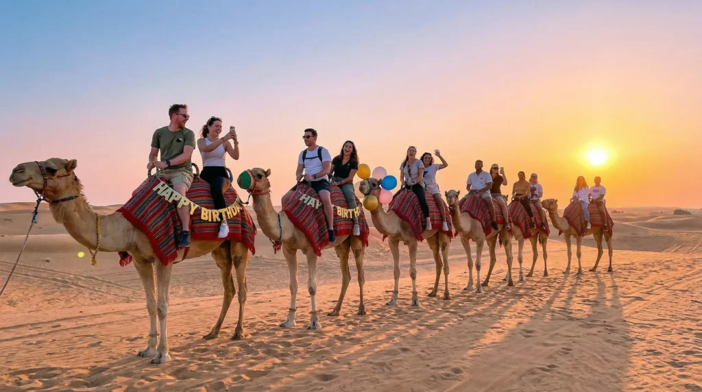 Group of friends enjoying a camel trek across the Dubai desert at sunrise, with golden sand dunes in the background.